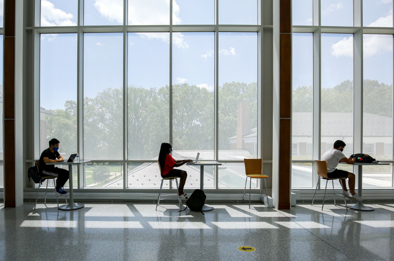 three students sit at desks in front of window